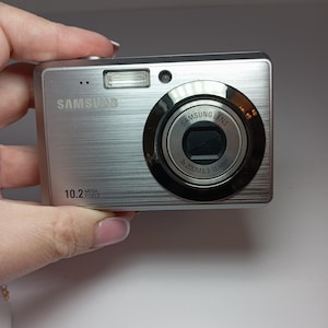 Może przedstawiać: Srebrny aparat cyfrowy Samsung z obiektywem z 3-krotnym zoomem. Aparat posiada wbudowaną lampę błyskową i napis "10.2 Mega Pixels". Obiektyw ma napis "Samsung Lens 3x Zoom 6.3-18.9mm".