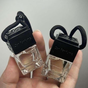 May include: Two black car air fresheners with black string hangers. The air fresheners are square shaped and have a clear glass bottle with a liquid inside. The text on the air fresheners reads "Car Perfume" and "New Perfume".