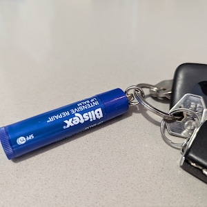 Puede incluir: Un tubo de bálsamo labial azul con el texto "Blistex Intensive Repair Lip Balm SPF 15". El bálsamo labial está unido a un llavero plateado con un llavero negro.