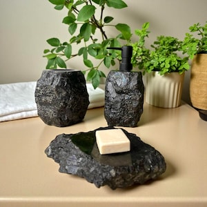 以下が含まれることがあります： 黒い石のバスルームセット。石鹸ディスペンサー、タンブラー、石鹸皿が含まれています。ディスペンサーは黒いポンプ付きで、石鹸皿には石鹸が置かれています。セットは明るい色の表面に置かれ、背景には植物があります。