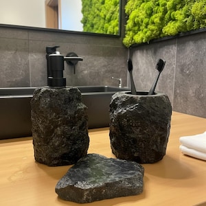 May include: A black stone bathroom set including a soap dispenser, toothbrush holder with two black toothbrushes, and a soap dish. The set is made of dark gray stone with a rough, textured surface. The items are displayed on a light wood surface.