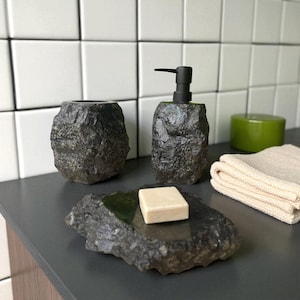 May include: A bathroom set made from dark grey, textured stone. It includes a soap dispenser with a black pump, a tumbler, and a soap dish holding a bar of soap. A green container and a folded beige towel are also visible.
