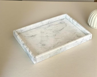 May include: Rectangular marble tray with a white and gray veined pattern. The tray has a raised edge and is designed to hold small items. The tray is made of natural stone and has a smooth surface.