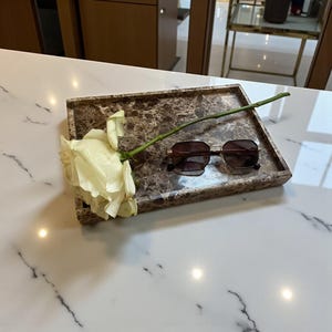 May include: A rectangular marble tray with a brown and white pattern holds a pair of sunglasses and a white rose. The sunglasses have a gold frame and dark lenses. The rose is light yellow.