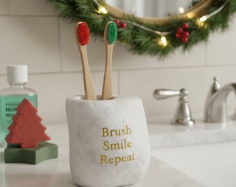 May include: A white marble toothbrush holder with gold lettering reading "Brush Smile Repeat". Two toothbrushes with red and green bristles are placed inside. A Christmas wreath and a red tree-shaped soap are in the background.