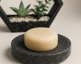 May include: A light beige, round bar of soap rests on a dark gray, circular soap dish. The soap dish has a textured surface. In the background, a black geometric planter holds small plants and white pebbles.