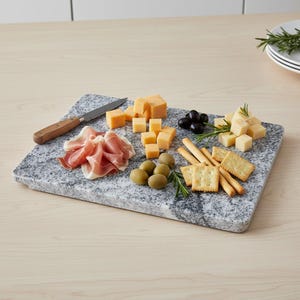May include: A rectangular gray granite serving board with various food items. Includes cubes of cheese, olives, crackers, breadsticks, prosciutto, and a knife with a wooden handle. Rosemary sprigs add a decorative touch.
