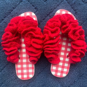 Könnte beinhalten: Ein Paar rot-weiß karierte Flip-Flops mit einem roten, gerüschten Häkeldesign. Die Sandalen werden auf einer blauen, strukturierten Oberfläche präsentiert und zeigen eine handgefertigte, sommerliche Ästhetik.