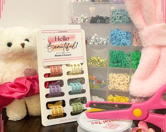 May include: A set of colorful hair clips in a clear plastic packaging with the text "Hello Beautiful! Mark & Lola's" on the front. The packaging is next to a container of colorful beads, a pink fuzzy headband, a pink and yellow pair of scissors, and a white teddy bear wearing a pink satin bow.