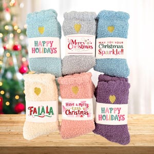 May include: Assortment of fuzzy socks in various colors, including blue, gray, and pink. Each pair features a gold heart and a band with holiday greetings like "Happy Holidays" and "Merry Christmas."