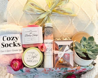 May include: A gift box filled with self-care items, including a white blanket, pink socks, a candle that says "Your light is so bright", a bar of soap, bath salts, and a small potted succulent.
