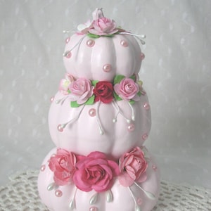 May include: A stacked pumpkin decoration painted light pink, featuring three tiers adorned with artificial pink and red roses, green leaves, and scattered round pearl-like beads. The arrangement is topped with a single small pink rose.