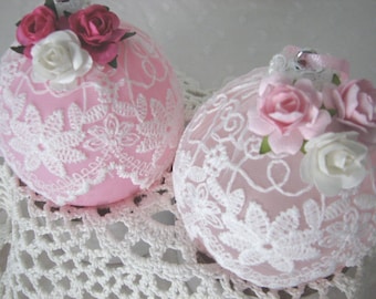 May include: Two spherical ornaments in light pink, each decorated with white lace overlays featuring floral patterns. The tops of the ornaments are adorned with clusters of small artificial roses in shades of pink and white, attached to silver loops.