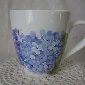 May include: White ceramic mug with a hand-painted design of purple and blue hydrangeas. The mug has a rounded shape and a handle.