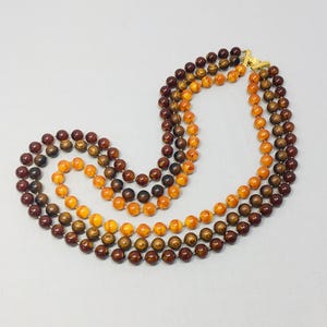 May include: Three-strand necklace featuring round beads in shades of brown and orange. The beads are strung together, creating a layered effect. The necklace has a gold-colored clasp.