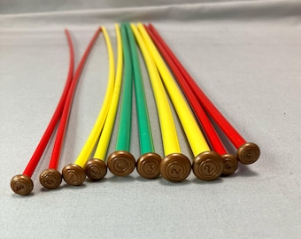 May include: A collection of colorful knitting needles in red, yellow, and green. Each needle has a brown, circular end with a number. The needles are arranged in a fan shape, with the tips pointing towards the viewer.