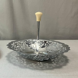 May include: Silver-toned serving dish with a decorative floral design. The dish has a handle topped with a cream-colored knob. The rim of the dish has a scalloped edge.