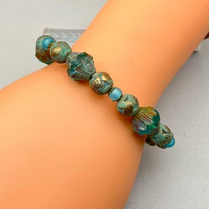 May include: A beaded bracelet featuring turquoise and gold-colored beads. The bracelet has a mix of round and uniquely shaped beads, with a vintage or antique aesthetic. The beads are strung together, creating a decorative accessory.