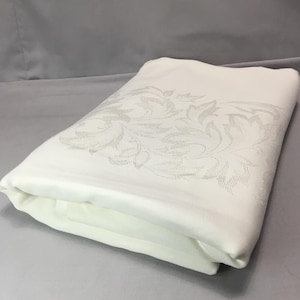 May include: Folded white tablecloth with a detailed floral pattern in a lighter shade. The fabric appears to be a smooth, possibly satin or linen, with a subtle sheen. The tablecloth is neatly folded, suggesting it's ready for use.