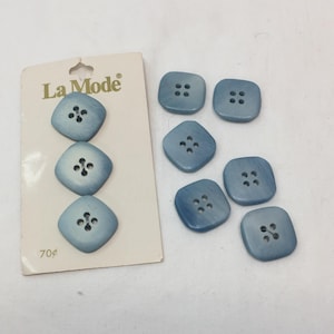 Puede incluir: Un conjunto de botones cuadrados azul claro con cuatro agujeros, algunos aún adheridos a una tarjeta de la marca "La Mode". Los botones tienen un aspecto ligeramente texturizado y una forma cuadrada redondeada. La tarjeta tiene el precio de 70¢.