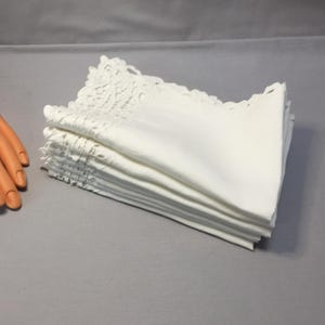 Puede incluir: Una pila de servilletas de lino blanco con delicados bordes de encaje. Las servilletas están cuidadosamente dobladas y apiladas, mostrando su aspecto limpio y nítido. El encaje añade un toque de elegancia al diseño sencillo.