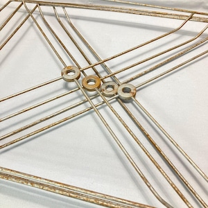 Könnte beinhalten: Ein antiker, geometrischer Metallrahmen mit einer verwitterten, cremefarbenen Oberfläche und sichtbarem Rost. Der Rahmen weist sich kreuzende Linien und kleine, kreisförmige Unterlegscheiben in der Mitte auf, was auf ein Vintage- oder Industriedesign hindeutet. Der Gesamteindruck ist gealtert.