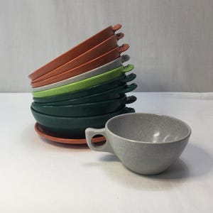 May include: A stack of round ceramic bowls in shades of terracotta, light gray, lime green, and dark forest green, each featuring a small side handle. Beside the stack sits a single light gray ceramic cup with a matching side handle design.