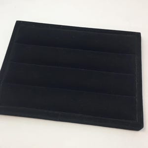 May include: A black velvet jewelry display tray with four horizontal slots. The tray is rectangular and has a slightly raised edge. The surface is soft and plush, designed to hold and showcase jewelry items.