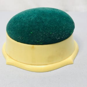 May include: A vintage pin cushion. It has a dark green, plush top and a light yellow, scalloped base. The base has a ribbed design and a curved edge. This sewing accessory is designed to hold pins.
