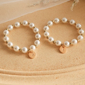 Op de afbeelding: Twee parelarmbanden met gouden accenten en een gouden bedel met de letter "E". De armbanden zijn gemaakt van witte parels en kleine gouden kralen. De ronde bedel hangt aan de armband.