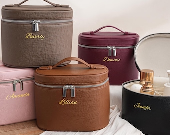 May include: Five oval-shaped cosmetic cases with top handles and silver zippers. The cases are colored taupe, pink, burgundy, brown, and black, each featuring a gold embroidered name: Beverly, Amanda, Dominic, Lillian, and Jennifer.