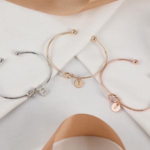 May include: Three gold, silver, and rose gold knot bangle bracelets with initial charms. The bracelets are on a white surface with a brown ribbon.