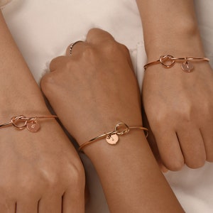 May include: Three gold knot bracelets with a small round charm on each. The charms have a single letter on them.