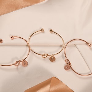 May include: Three gold-toned bracelets with a knot design and a small round charm. Each charm has a different letter on it.