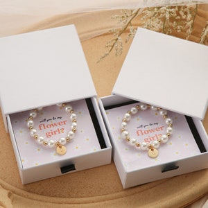 May include: Two white gift boxes with lids open, each containing a pearl bracelet with a gold charm. The boxes have a floral design and the text "Will you be my flower girl?" printed on the inside.