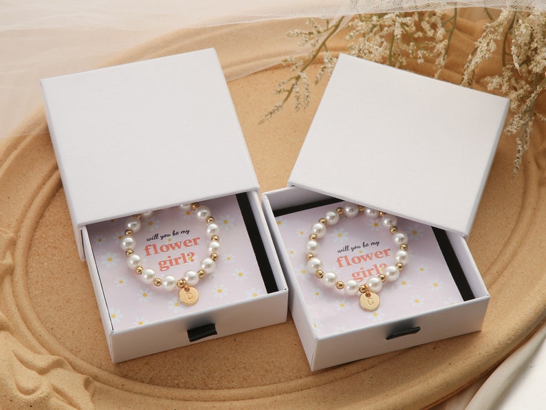 May include: Two white gift boxes with lids open, each containing a pearl bracelet with a gold charm. The boxes have a floral design and the text "Will you be my flower girl?" printed on the inside.