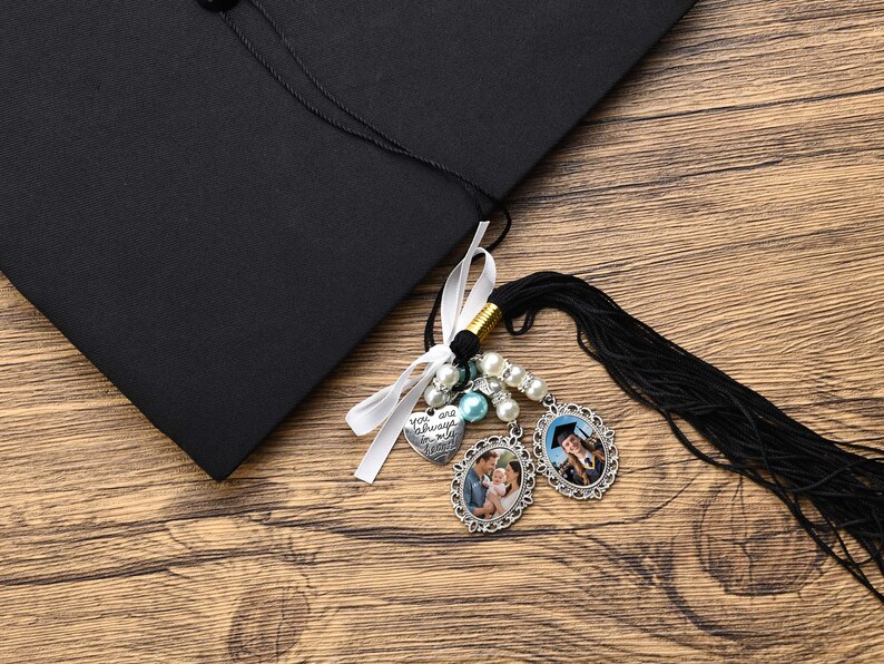 May include: A black graduation cap with a tassel and charms. The charms include a heart with the text "You are always in my heart", two oval photo charms, and a string of pearls and blue beads.