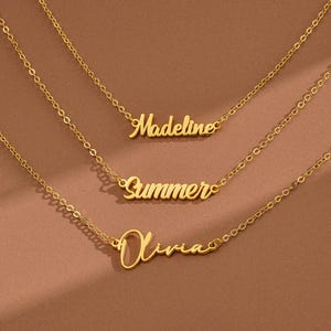 Könnte beinhalten: Drei goldfarbene Namensketten mit filigranen Kettengliedern. Die Namen "Madeline", "Summer" und "Olivia" sind in einer Schreibschrift dargestellt. Die Halsketten sind auf einem braunen Hintergrund angeordnet.