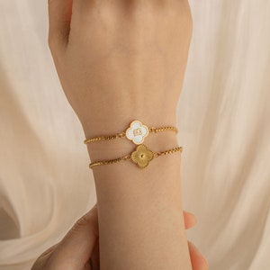 Puede incluir: Dos pulseras de oro con dijes en forma de trébol. El dije superior es blanco con una letra "B" dorada en el centro. El dije inferior es dorado con un diseño texturizado.