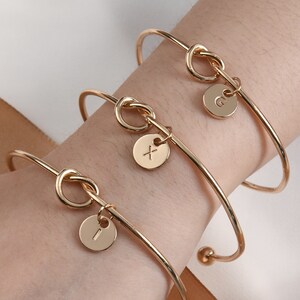 May include: Three gold knot bangle bracelets with initial charms. The charms are round and have the letters 'G', 'X', and 'I' engraved on them.