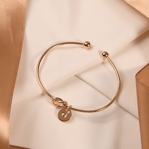 May include: A gold-toned bangle bracelet with a knotted design and a small round charm with the letter "O" engraved on it.