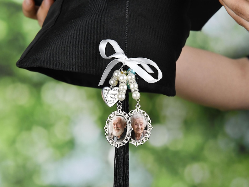 May include: A black graduation cap with a white ribbon and a charm attached. The charm is a silver heart with the words 'You are always in my heart' inscribed on it. The charm is attached to a string of white pearls and two silver oval frames with photos of older people inside. The frames are attached to a black tassel.