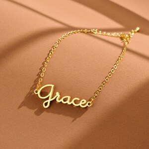 Pode incluir: Uma pulseira dourada com o nome "Grace" em escrita cursiva. A corrente fina está sobre uma superfície castanha. Esta joia personalizada é um acessório de moda.