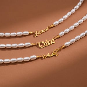 May include: Three pearl necklaces with gold name pendants. The names "Lillian", "Chloe", and "Aisha" are displayed in a delicate script. The necklaces are set against a warm brown background.
