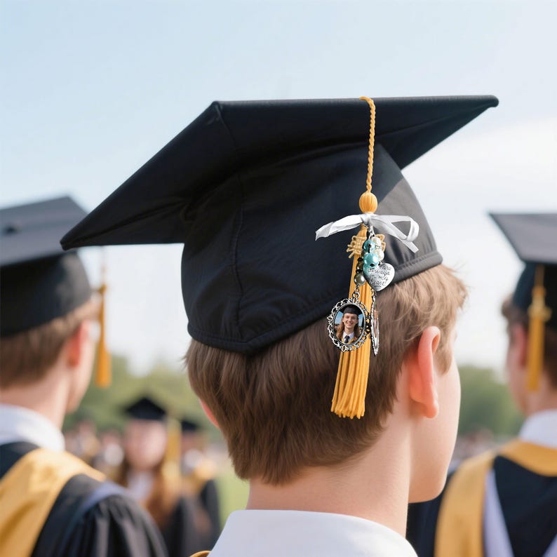 May include: A black graduation cap with a gold tassel and a charm attached to the tassel. The charm features a small photo, a heart, and a bead.