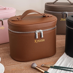 Könnte beinhalten: Personalisierte Kosmetiktaschen in Rosa, Braun, Grau und Schwarz. Jede Tasche hat einen Griff, einen Reißverschluss und einen Namen in goldfarbenen Buchstaben. Make-up-Pinsel sind im Vordergrund zu sehen.