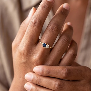 May include: A gold ring featuring a pear-shaped, dark blue gemstone and a pear-shaped, clear gemstone. The ring also has a small, round, clear gemstone. The ring is on a finger.