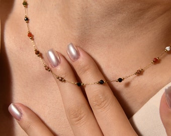 May include: A gold chain necklace with small, colorful beads. The beads are a mix of black, brown, red, and green. The necklace is on a person's neck.