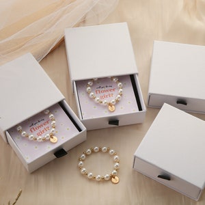 May include: Three white boxes with drawers, each containing a pearl bracelet with a gold charm. The bracelets are in small boxes with a pink background and white daisies. The text on the boxes reads "Will you be my flower girl?"