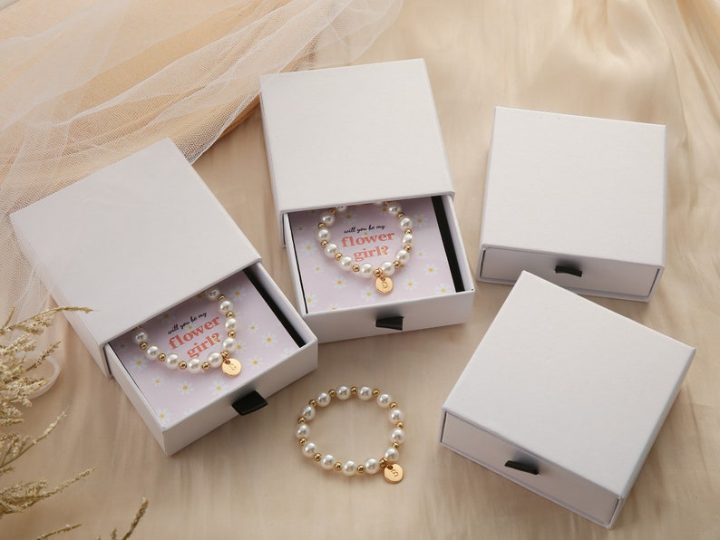 May include: Three white boxes with drawers, each containing a pearl bracelet with a gold charm. The bracelets are in small boxes with a pink background and white daisies. The text on the boxes reads "Will you be my flower girl?"